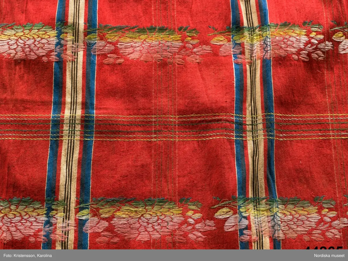 Kvadratiskt halskläde av  halvsiden.  Storrutig  på klarröd botten i satin  med  ränder i schatterande blått, beige, rött och brunt och med jacquardvävda tvärgående blombårder  i rosa, grönt  och gult silke med smala gula  kantbårder. Runtom upprispad kort frans. I ena hörnet en rund svart stämpel med svårläst text: Stockh. Det är Stockholms hallstämpel jfr. likadan t halskläde 44.265 som har en tydligare stämpel  från 1830-talet.
I mycket gott skick.
Troligen en brudschal. Se samma mönster i mörkblått 91.624 från Unnaryd i Småland.
/Berit Eldvik 2012-01-27