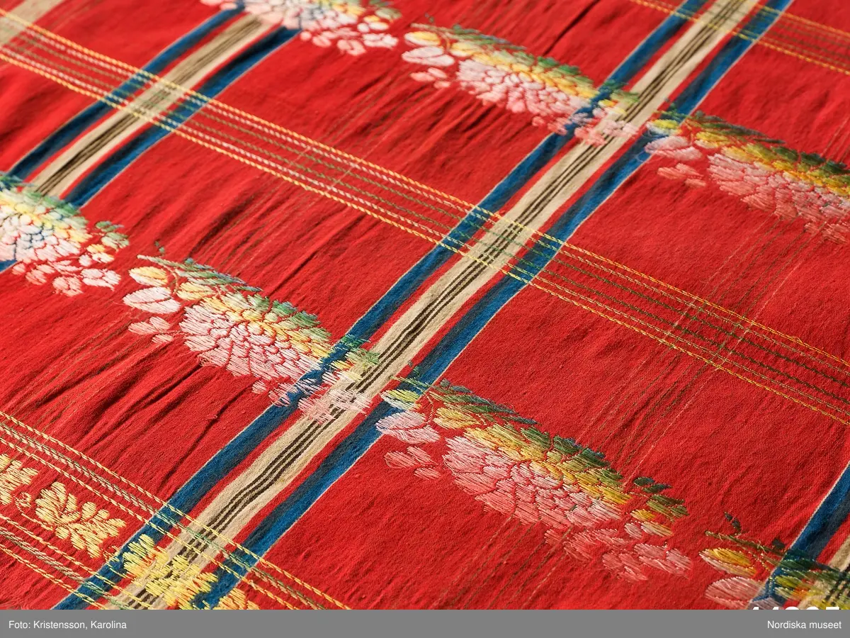 Kvadratiskt halskläde av  halvsiden.  Storrutig  på klarröd botten i satin  med  ränder i schatterande blått, beige, rött och brunt och med jacquardvävda tvärgående blombårder  i rosa, grönt  och gult silke med smala gula  kantbårder. Runtom upprispad kort frans. I ena hörnet en rund svart stämpel med svårläst text: Stockh. Det är Stockholms hallstämpel jfr. likadan t halskläde 44.265 som har en tydligare stämpel  från 1830-talet.
I mycket gott skick.
Troligen en brudschal. Se samma mönster i mörkblått 91.624 från Unnaryd i Småland.
/Berit Eldvik 2012-01-27