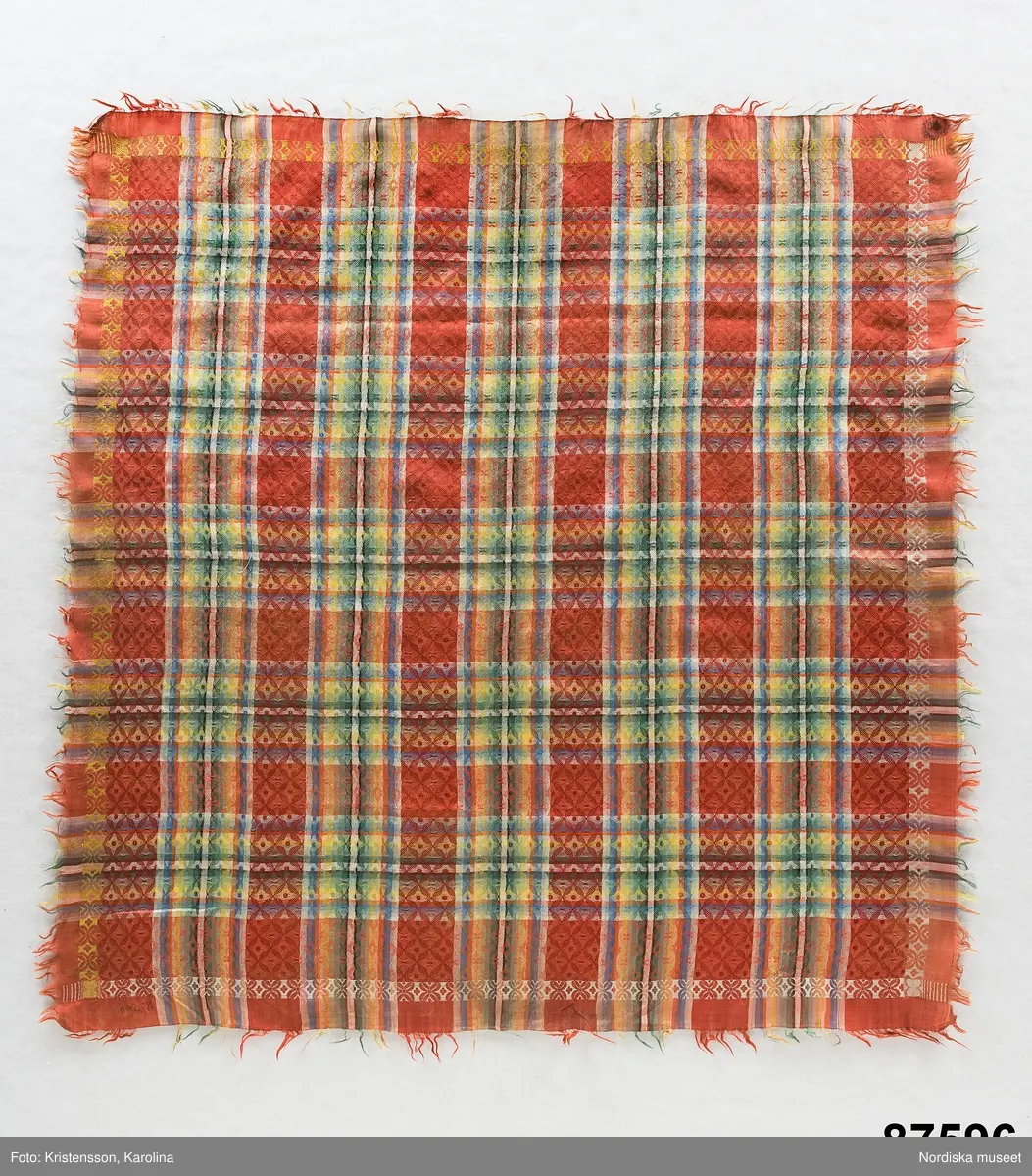 Kvadratiskt halskläde av siden, orangeröd botten med rutning i gult, grönt, ljusblått och vitt samt en smal ytterbård med stiliserad blomma längs 2 sidor i vitt och längs de andra i gult. Botten mönstervävd i ett bindemönstermönster med kryss och små stjärnor, i ytterkant utanför bården slät tuskaft. Uppsrispad kort frans runt alla sidor. I ena hörnet en rund svart hallstämpel med texten. Stockholm 1825 och i mitten St. Eriksbild. Troligen vävd i en skaftvävstol.
Typen var vanlig på 1820-talet och finns från flera håll i landet bl.a 98530 från Äppelbo och 93669 från Malung  i Dalarna. även 44279 från Västmanland och 91632, 152104, 162623 från Småland.
I fint skick
/Berit Eldvik 2011-11-23