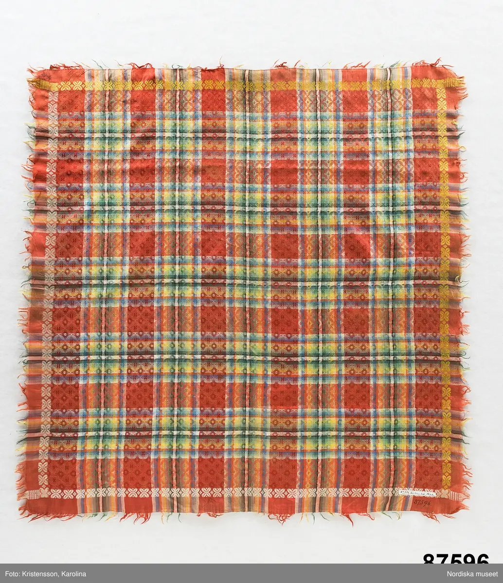 Kvadratiskt halskläde av siden, orangeröd botten med rutning i gult, grönt, ljusblått och vitt samt en smal ytterbård med stiliserad blomma längs 2 sidor i vitt och längs de andra i gult. Botten mönstervävd i ett bindemönstermönster med kryss och små stjärnor, i ytterkant utanför bården slät tuskaft. Uppsrispad kort frans runt alla sidor. I ena hörnet en rund svart hallstämpel med texten. Stockholm 1825 och i mitten St. Eriksbild. Troligen vävd i en skaftvävstol.
Typen var vanlig på 1820-talet och finns från flera håll i landet bl.a 98530 från Äppelbo och 93669 från Malung  i Dalarna. även 44279 från Västmanland och 91632, 152104, 162623 från Småland.
I fint skick
/Berit Eldvik 2011-11-23