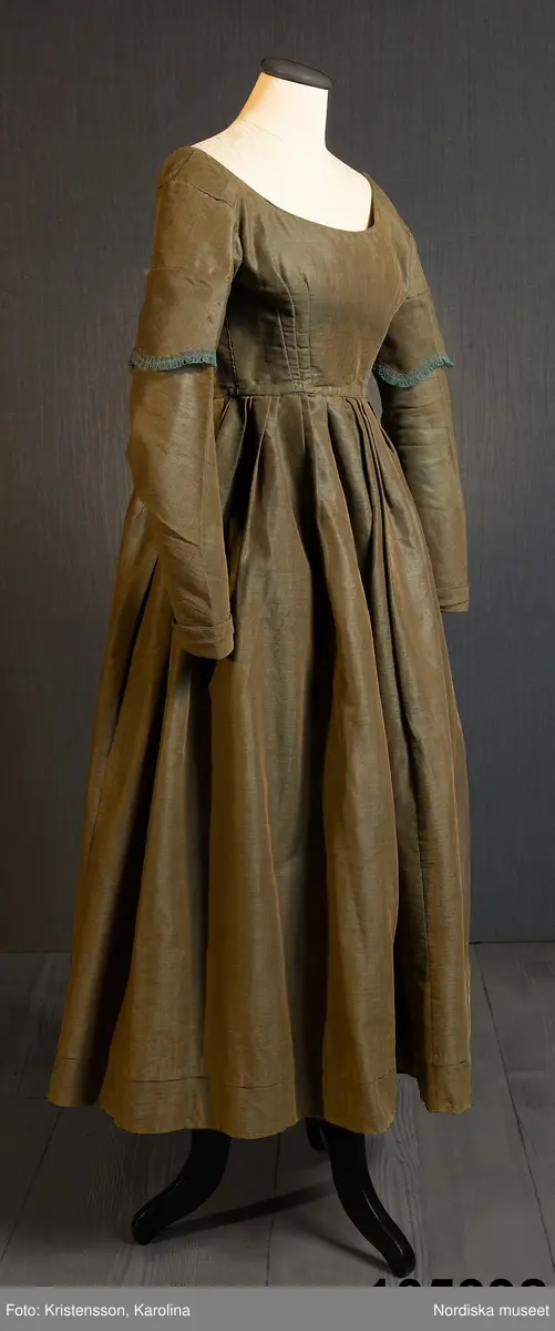 Hel klänning i 1830-talets modesnitt, av fint halvylletyg i kypert med varp av ljusbrunt bomullsgarn och inslag av grönt tunt glansigt redgarn som ger en grönskimrande yta. Livet går ned nästan till midjehöjd, figursytt med 2 framstycken med bakåtflyttade sidsömmar, ett ryggstycke med 6 inprovningar fördelade i två grupper. Vid båtringad halsringning, knäppning med tätt sittande hakar och hyskor. Isydda långa ärmar svängda med 2 sömmar  med 3 veck vid armbågen. Ärmsprund knäppt med hyska och hake samt 4 cm uppslag med rundade hörn. Upptill på ärmen besättning av tyget med stora rundade uddar kantade med klargrön bomullsfrans.  Passepoaler av tyget i alla sömmar. Livet fodrat med skotskrutig bomullslärft i rött, grönt, gult och vitt och i ärmarna ett smalrandigt bomullstyg i blått och rosa. Kjolen sydd av  5 våder, lagda veck runtom i midjan. sprund mitt fram. Uppsytt veck  en decimeter från fållen. Skoning med brun bomullslärft. Handsydd.
Har burits 1838 som ungmorsklänning, alltså av bruden dagen efter bröllopet, av Maria Margareta Mårtensdotter från Kysings, Vall sn på Gotland. Möjligen tillsammans med sidenhalsklädet 105385.
Anm. I  gott skick.
OBS Klänningen sitter bakochfram på fotona, knäppningen är fram.
/Berit Eldvik 2011-06-13