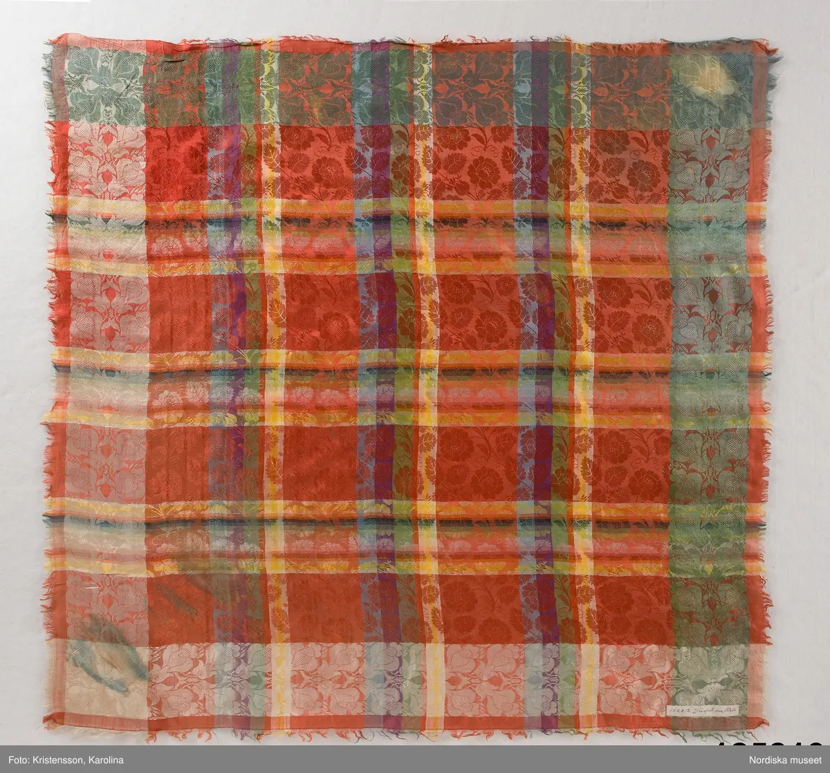 Kvadratiskt halskläde av siden, klarröd botten med rutor i grönt, ljusblått, ljuslila och gult med ett yttäckande invävt mönster, troligen med jacquard, med slingrande rosor. Kantbård med "päronlika" blommor/frukter som på 2 mötande sidor är gröna och på de 2 andra vita. Upprispad kort frans på alla sidor.
I ena hörnet en tydlig svart rund stämpel med texten: Stockh. hallstemp 1839.
Anm. Stor fläck i två av hörnen.
/Berit Eldvik 2011-11-11