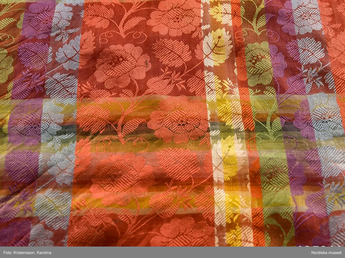 Kvadratiskt halskläde av siden, klarröd botten med rutor i grönt, ljusblått, ljuslila och gult med ett yttäckande invävt mönster, troligen med jacquard, med slingrande rosor. Kantbård med "päronlika" blommor/frukter som på 2 mötande sidor är gröna och på de 2 andra vita. Upprispad kort frans på alla sidor.
I ena hörnet en tydlig svart rund stämpel med texten: Stockh. hallstemp 1839.
Anm. Stor fläck i två av hörnen.
/Berit Eldvik 2011-11-11