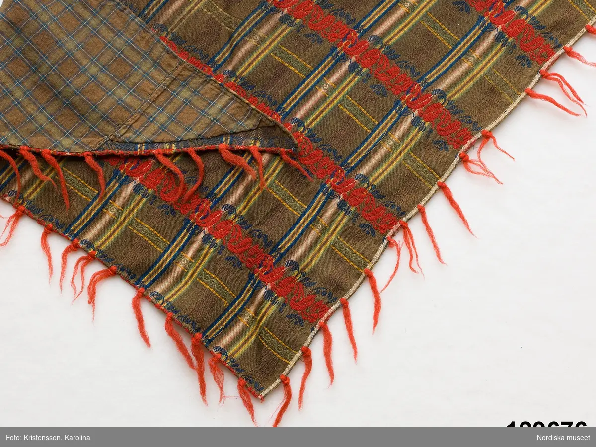 Trekantigt halskläde av troligen jacquardmönstrad bomullssatin.  Grönbrun botten randig i  ljusare grönt, gult och blått med invävd bladslinga i bottenfärgen. Tvärgående mönsterbårder i rött och blått  samt smalare i gult och grönt, vilket ger ett rutigt  tyg. Tvärbårderna i inslagsflotteringar.   Tyget har 2 materialskarvar. I ytterkanterna iknuten gles frans av rött ullgarn.Fodrad till större delen med ett grön/brunrutigt bomullstyg, snibben bak ofodrad.
 Tillverkad av delar från ett kvadratiskt halskläde.
/Berit Eldvik 2012-02-09