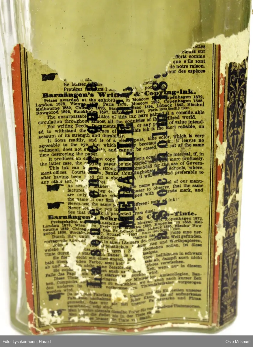 Flasken har etikett over tre sider. Den har mørk beige bunnfarge, og trykk i sort, blått, rødt og gull. Tegning av en bjørn som rører i en bøtte med blekk under teksten "FABRIKSMÄRKE".