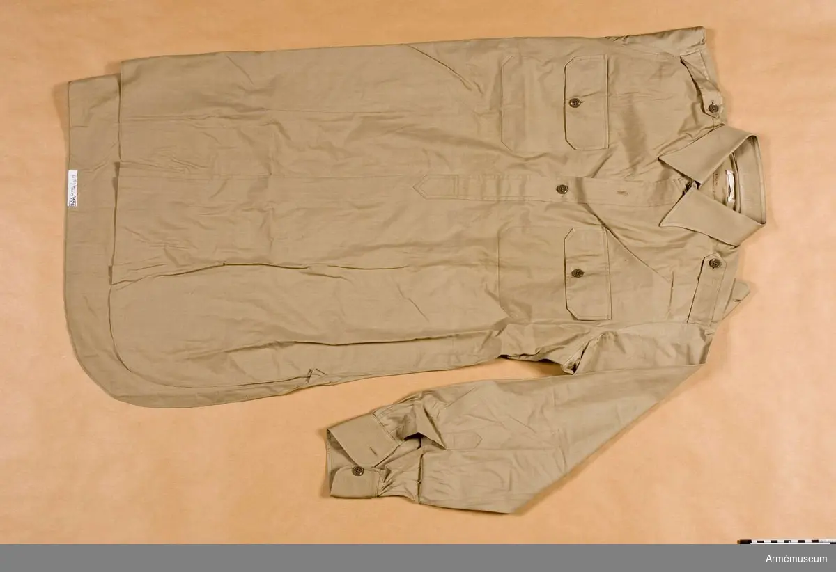 Av brunbeige bomull. 460 mm lång öppning framtill som knäppes med tre knappar. Skjortan har nedvikt krage. Två bröstfickor med lock och knapp. Axelhylsor.
(NK står vanligen för Nordiska Kompaniet, 19830207 BoM.)