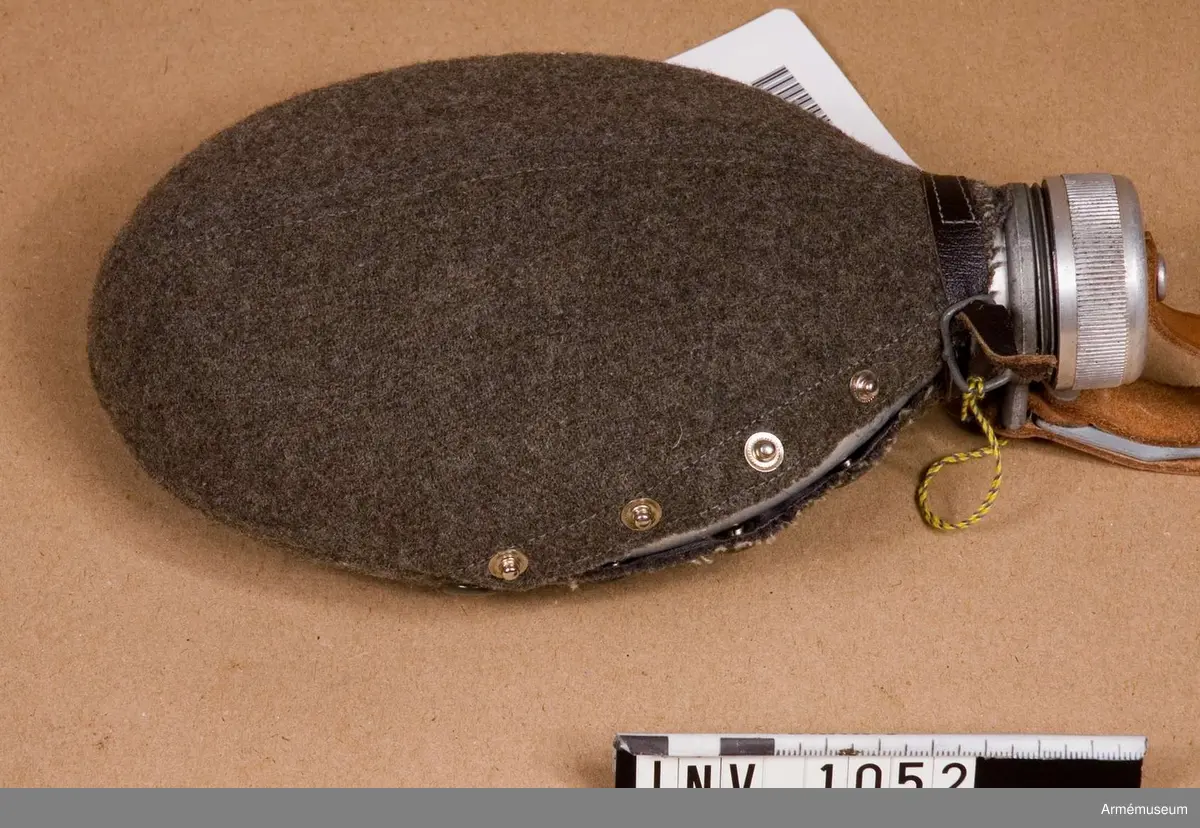 Samhörande nr är 1017-1059, 1062-1063, 1072-1073.Fodral t dricksflaska m/1929.Av kommisskläde, m/1929. Övre delen, utsidan, är försedd med  skarv på mitten med nedstickade sömmar. Ett sprund med fyra  tryckknappar. Undre delen är något mindre tillskuren med skarv  på mitten och nedstickade kanter. På undre stycket sitter fyr tryckknappsöverdelar som är överklädda med gröngrå celluloid. Över- och underdel är hopsydda med en söm till sprundet. Runt  "halsen" hålls fodralet till med en läderrem som knäpps med  litet grått metallspänne. Fodralet förvaras påträtt över  flaskan. Se AM 1051.Har tillhört mobiliseringsutrustning vid Armémuseum. 