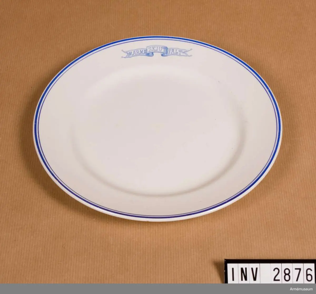 Vitt porslin med blå dekor; runt ytterkanten en något kraftigare rand än den innanför. Emblem i form av banderoll med text "MARMA SKJUTFÄLT". Undersidan märkt "Gustavsberg 1718 G5". Har använts vid off.mässen, Marma läger. Porslinet har tillh. A 1 och har förmodligen anskaffats i början av 1930-talet som ersättning för porslin som blev förstört när mässen brann ner feb 1930.

Samhörande nr är 2625-2630, 2876-2881 (2876-2881).