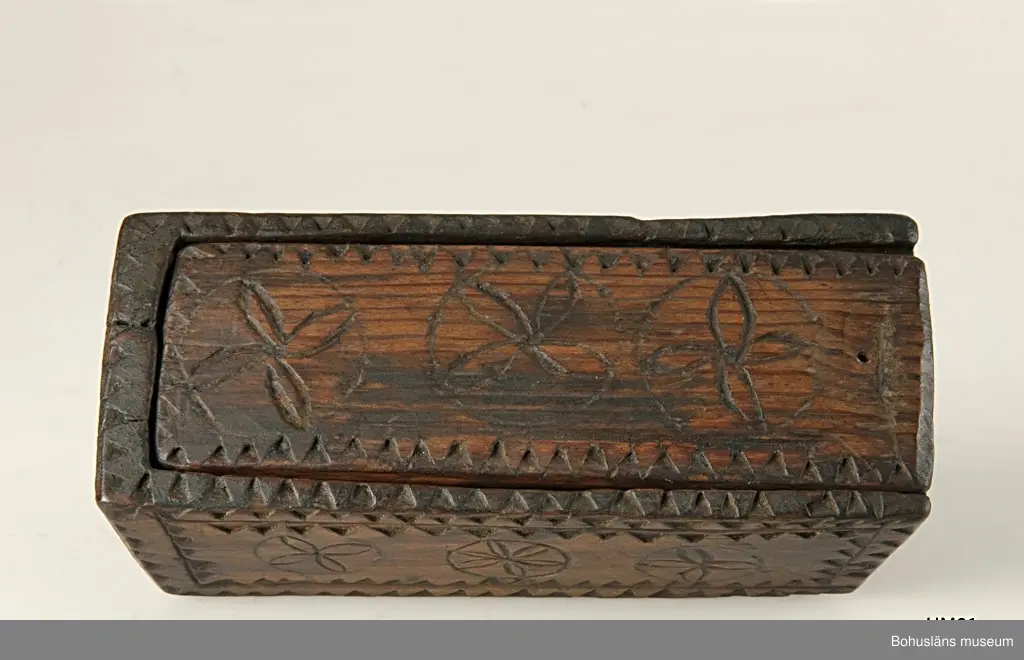 Rektangulär låda med skjutlock och något snedställda kortsidor.
Skuren dekor bestående av rosetter, kors samt uddsnitt längs kanterna.
På en av långsidorna är årtalet "1848" och initialerna "AN" inskurna.
Inuti ligger en äldre etikett från ca sekelskiftet.
Kan vara en friargåva.

Litteratur:
Gjaerder, Per, Esker og tiner, C. Huitfeldts förlag, Oslo, 1981, s. 41.
Knutsson, Johan, Friargåvor, Nordiska museets förlag, Stockholm, 1996.

Ur handskrivna katalogen 1957-1958:
"Trälåda
1870-1887
Bottenmått: 14 x 5,5 H. 5. Trä med skuren ornering. Föremålet helt. Bohusl."

Lappkatalog: 84