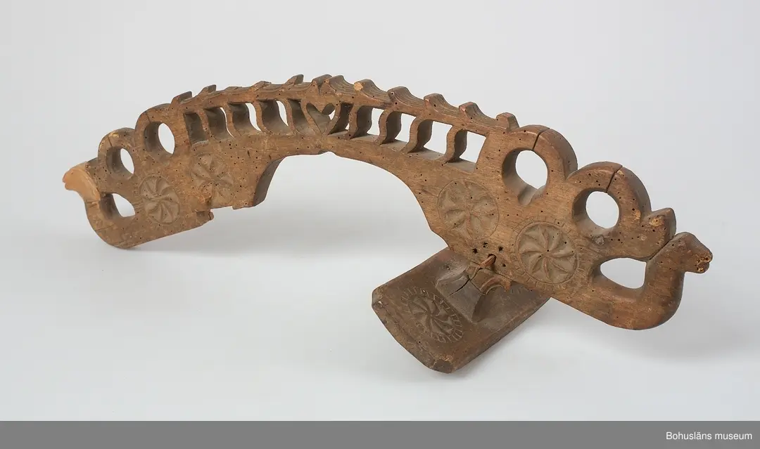 Böjd, genombruten båge med karvsnittsdekor på båda sidor bestående av rosetter. Längst ut på vardera ända är ett drakhuvud.
Rektangulära fästen, vilka vilar på hästens manke. Karvsnittsdekor med rosetter på en av dem. Den andra är sekundär och har skurna cirklar samt är defekt.
Ett drakhuvud är sekundärt.
Skadedjursangrepp.

Litteratur: Edvardsson, Ingmar, Rack, ryss och rissla, Stockholm, 1995, 
s. 66 samt bild s. 42.
Kjellberg, Sven T.: Svenska selkrokar av trä i Nordiska museet. Fataburen 1926. 
Nylén, Anna-Maja, Hemslöjd, Håkan Ohlssons förlag, Lund, 1978, s. 396-401.

Ur handskrivna katalogen 1957-1958:
Selranke av trä
I 3 delar a-c, a) = oket L. 59 cm; b) "stödplatta", L. 19,5 cm; c) d:o, L. 20 cm (nygjord). Oket har snidad dekor i genombrutet arbete m. drakhuvud i ändarna (ett nygjort); hela hela selranken har karvsnittsdekor. Omålad. Maskäten. 

Lappkatalog: 40
Hugo Hallgren var son till salteriidkaren och handelsmannen Johan Jacob Hallgren och Emelia Gerle. Företaget började sälja brissling på 1850-talet och 1864 startade man konservfabrik på Gullholmen som senare döptes till J. J. Hallgren & Söner. Sönerna var Gustav Richard, f. 1858 och Oscar Hugo, f. 1867.  Här introducerades flera nyheter inom ansjovisindustrin,  bl a Sveriges första skinn- och benfria delikatessill. Företaget var också först med att lägga in ansjovis i hermetiskt slutna kärl av bleckplåt. 1898 köptes Hallgrens konservfabrik upp av det nybildade storföretaget AB Sveriges Förenade Konservfabriker (SFK). 1901 flyttade Hugo Hallgren till Ellös där han startade en ny konservfabrik.



För uppgifter om förvärvet, se UM5155.