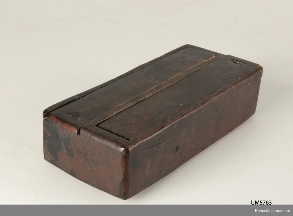 Rektangulär låda, uppdelad i två parallella fack med varsitt skjutlock upptill.
Målat med mörkt röd färg.
Inuti ett av facken finns fragment av fnöske.

Litt.; Gjaerder, Per, Esker og tiner, C. Huitfeldts förlag, Oslo, 1981, s. 48-50.

Ur handskrivna katalogen 1957-1958: 
Trälåda m. två skjutlock
a) L. 22,5 Br. 9,7, H. 5,3 cm, rödmålad; m. 2 fack, under m. ett skjutlock b) och c); deras L. 21,2 cm, Br 4 cm.

Lappkatalog: 84