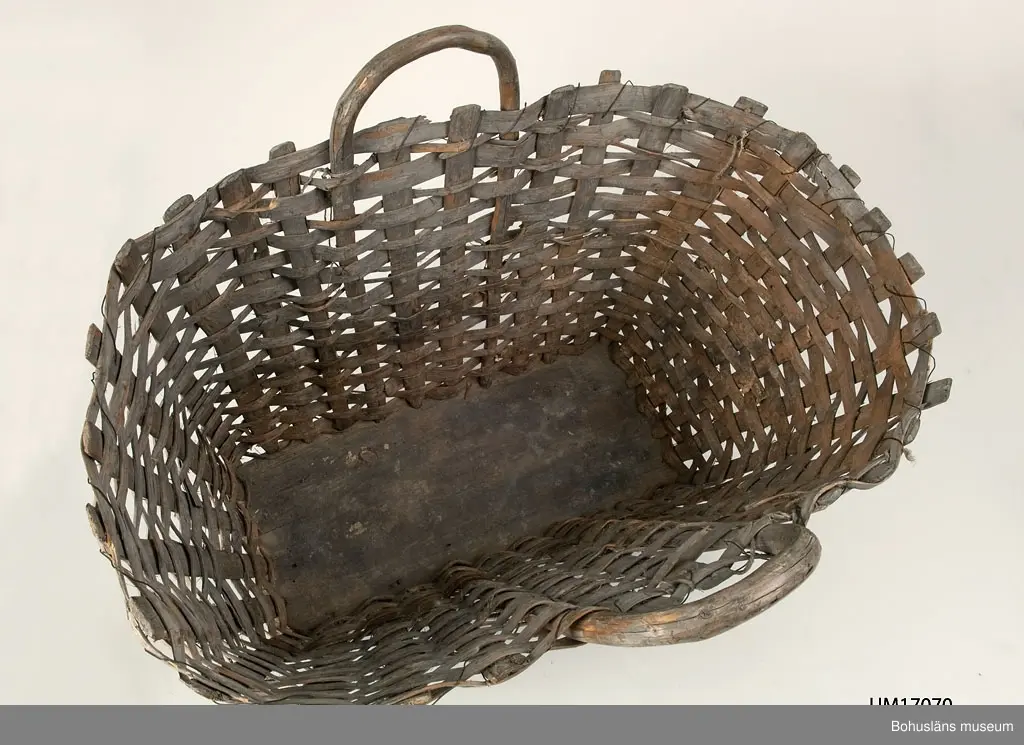 Oval korg med rektangulär bottenplatta. På varje långsida är det ett bärhandtag av en böjd gren. En lindad järntråd förstärker kanten upptill.
Troligen använd till rotfrukter under 1900-talets första hälft.