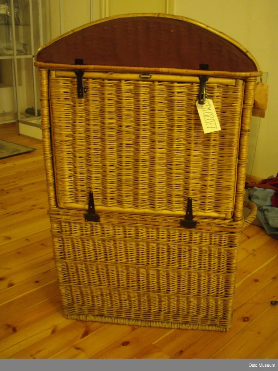 Reisegarderobe, amerikakoffert brukt til ballettutstyr i rotting med buet og lerret-trukket tak. 2 bærehanker. Åpning i front med beslag av metall. Innvendig trukket med ullstoff (ullteppe).