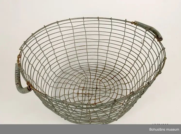 Rund korg vilken är svagt konande nedåt. Två ledade bärhandtag omvirade med ståltråd.
Korroderade.
Mått på den andra korgen: L 53,5, B 55, H 22 cm