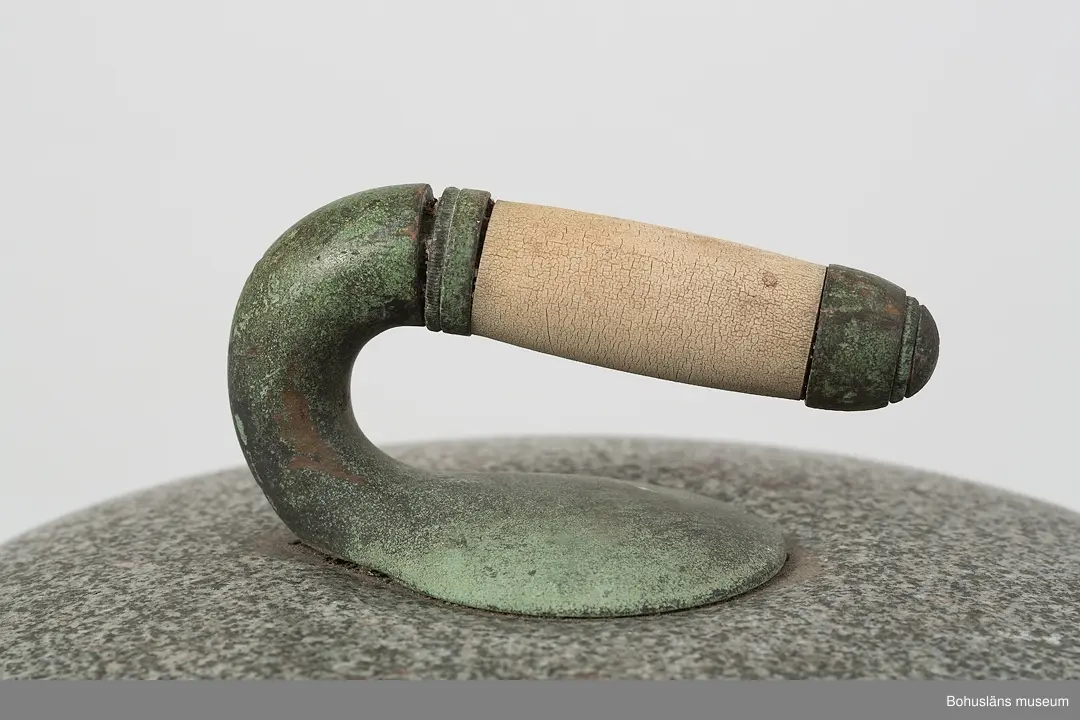 Cirkelrund curlingsten med böjd handtagsdel av dekorgjuten brons med grepp av trä. Stenen är mycket jämn med slätpolerad ovan- och underdel och med ett råslipat mellanparti, 5 cm högt, som löper runt hela stenen. 
Handtaget är grönkorroderat och något stukat.