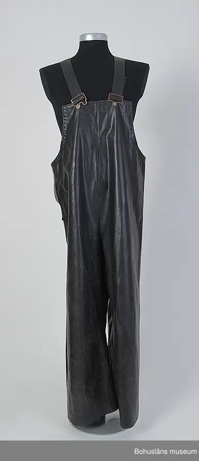 Hängslebyxor till oljeställ med jacka och sydväst. Plastbelagt flanelltyg med både svetsade och maskinsydda sömmar. Hängslen med metallknäppen - av olika utseende - baktill på ryggsidan ljusa resårer. Metallknappar med ingjuten beteckning "EWERTS".
Slits i ena sidan med tryckknapp.