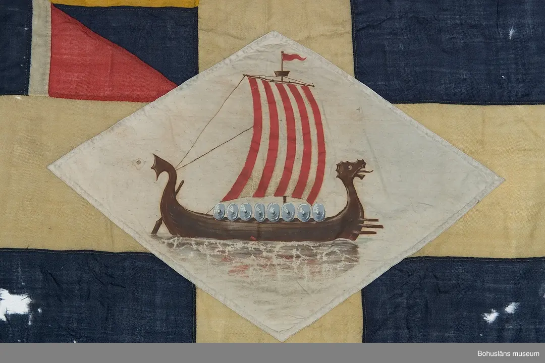 Segelsällskapet Vikens flagga, SS Viken. 
Svensk tvåtungad  flagga med parti "Silllsallaten", svensknorska unionsflaggan i övre hörnet samt infällt handmålat rombiskt emblem placerat mitt i det gula korset. På emblemet är målat ett vikingaskepp. 
Märkt "KASEN" med svart märkbläck. Hand- och maskinsömnad.
Skador. Eventuellt har flaggans form ändrats.