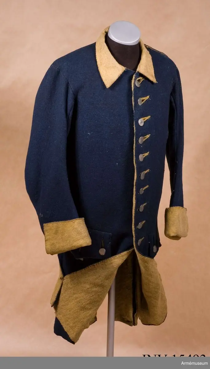 Rock av blått kläde med gul krage och ärmuppslag. Fodrad med gul boj i livet och med linne i ärmarna. Försedd med tennknappar.