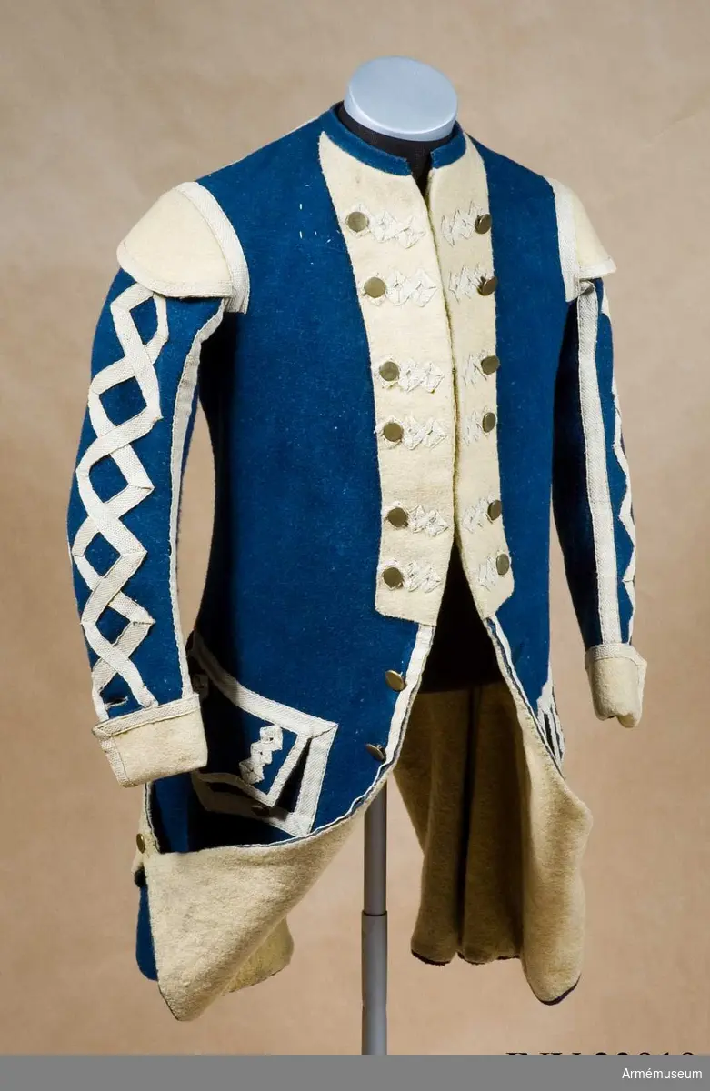 Rock med prydnader för spelet; mellanblå med gul rabatt (bröstrevärer), svalbon (axeldekoration) och ärmuppslag. Vit redgarnsdekor bruklig för spel. Rocken har sammanlagt 23 knappar. Knapparna i framkanten fastträdda med vitt band från avigsidan. En knapp i vardera skört (en ny). Två knappar på ficklocken (höger sida fattas en). Rocken är fodrad med gult ylletyg. Rocken ihopfästes med hyskar och hakar.