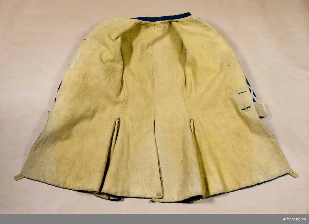 Rock med prydnader för spelet; mellanblå med gul rabatt (bröstrevärer), svalbon (axeldekoration) och ärmuppslag. Vit redgarnsdekor bruklig för spel. Rocken har sammanlagt 23 knappar. Knapparna i framkanten fastträdda med vitt band från avigsidan. En knapp i vardera skört (en ny). Två knappar på ficklocken (höger sida fattas en). Rocken är fodrad med gult ylletyg. Rocken ihopfästes med hyskar och hakar.