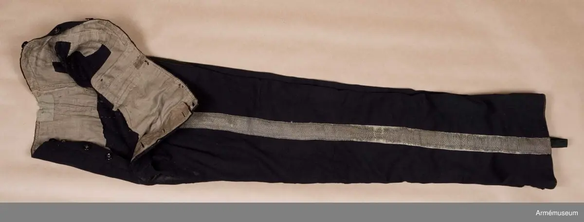 Mörkblå långbyxor med skinnskoning i baken och på benens insida, samt revärer av metallgalon i sidorna.

Byxorna är sydda av mörkblått kläde och har 4 cm breda revärer av silvergalon på benens utsidor. De är ursprungligen av en äldre modell med hel lucka fram, men har sytts om till gylf mitt fram, troligen i samband med att gylf fastställdes för långbyxor 1845. I varje sida syns spår efter luckan i form av ett smalt insnitt, samt ett igensytt knapphål.
Gylfen har dold knäppning med 4 enkla svartlackade knappar.
Byxorna är skinnskodda med brunt skinn i baken och på benens insidor, men det syns även rester efter en rad stygn ovanför knät på båda ben som eventuellt kan vara spår efter en äldre skinnskoning.
Byxorna är ofodrade, men har en infodring av oblekt bomullstyg i linningen upptill och en bit ner på framstycket. Infodringen är lappad på flera ställen.
På vänster byxben vid knät syns en stor lagad reva i tyget. Nertill vid fållen sitter i vänster byxben en sekundär hälla av svart resårband, samt en sekundär svart plastknapp. Dessa har troligen satts dit i samband med att byxorna varit utställda.
På insidan av höger bens fåll sitter två små knappar för fastsättning av hälla. En lapp eller lagning är dessutom iskarvad vid höger knä.
Byxorna har 3 knappar bak och 4 fram för fastsättning av hängslen. De två knapparna på höger sida fram är kullriga mässingsknappar, medan övriga är 4-hålsknappar av svartlackad metall.