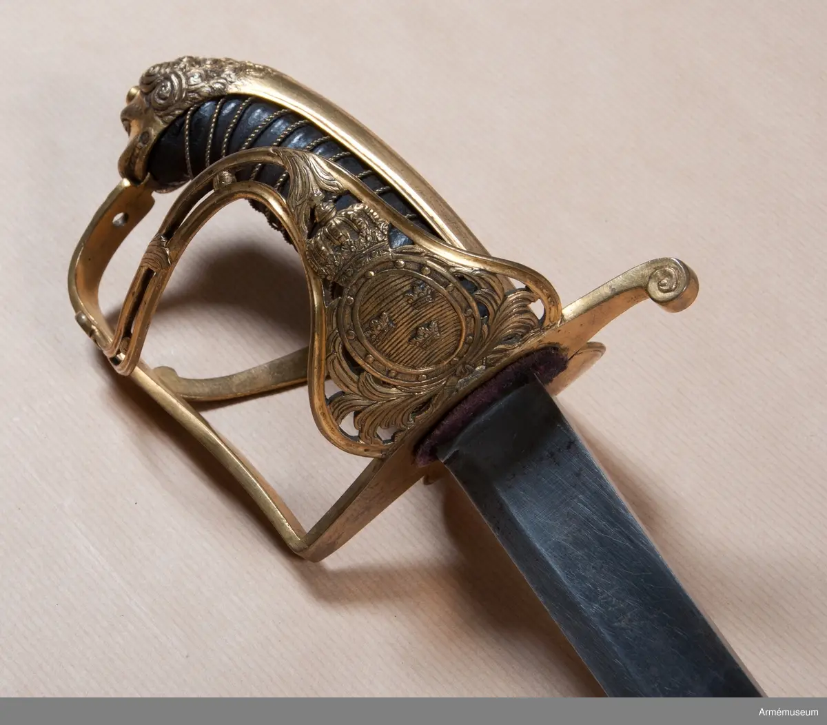 Klingans bredd upptill: 30 mm.Klingan är krökt, skålslipad och har nedtill en svart  betonad, tveegad klack. Fästet är av förgylld mässing. Kaveln är klädd med svart skinn en spiralräffla 16 varv. I bottnen på denna räffla är en av två parter hopsnodd, tunn mässingtråd anbringad. Baktill har kaveln ryggbeslag, som upptill övergår i en kappa, vilken är ciselerad i form av ett lejonhuvud och på vilken sitter en liten nitknapp. Parerstången är en rak och har fyrkantigt tvärsnitt samt av- slutas baktill av en nedböjd, på sidorna med ett spiralornament prydd knapp. Från parerstången främre ände utgår handbygeln i rät vinkel. Handbygeln är längs mitten litet försänkt och har längs var och en av framsidans kanter en ingraverad rand. Litet framför kappan genombrytes den av ett runt hål, och kring detta hål finns ciselerade ornament vilka bilda en stiliserad krans. På yttersidan finnes en uppåtböjd, delvis genombruten parerplåt, som är prydd med krönt sköld med tre kronor, vilken sköld omgives av ett par palmkvistar, allt i ciselerat arbete. Från parerplåtens spets utgår en delvis genombruten och sparsamt med ciselerade ornament prydd S-formig spång, som går till hand- bygelns övre del. På innersidan finns en liknande men mindre, ej genombruten och ej ciselerad parerplåt, från vars spets en S-formig spång går till handbygeln, vilken den träffar något längre ned än ytter- spången. Innerspången prydes längs kanterna av ingraverad rand.