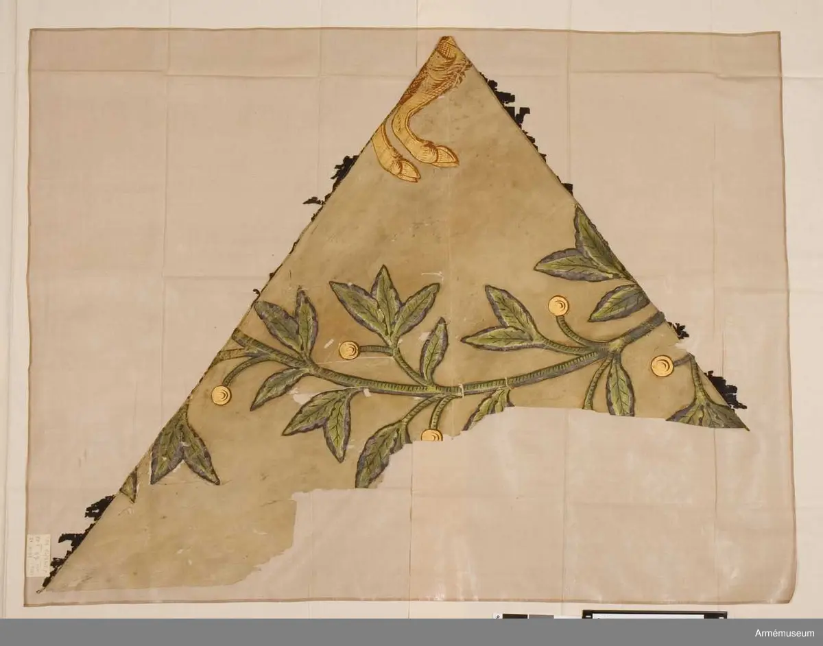 Trekantigt fragment av fana av gulnat siden med bemålning i form av en grön olivgren (?) och Hälsingebockens framben i guld och rött. De saknade fälten i fanan har troligen varit svarta.