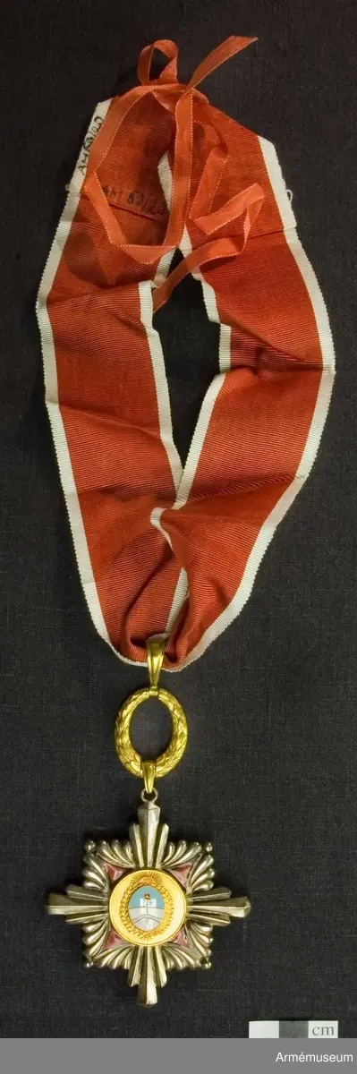 Kommendörstecken av argentinsk förtjänsteorden. På framsidan en man hållande argentinas fana i blått och vitt med texten "AL MERITO". På baksidan vapenskölden för Argentinas president.

Med band i rött med vita kanter.