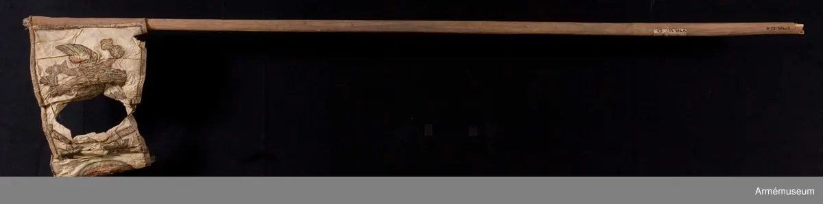 Duk av vit sidentaft och strumpa av rött linne. Motivet målat i guld- och silver samt rött och grönt bestående av en krönt örn, måne, sol och stjärnor, På duken en pappersetikett som anger att den tagits i slaget vid Saladen 1703.
