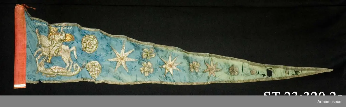 Entungad lansfana. Duk av blå kinesisk sidendamast med målad dekor. I 1703 års inventarium anges motivet vara St Göran men enligt den ryska inskriptionen är det Feodor Stratilat . Rosor och stjärnor av silver. Duken kantad med en silverbård. Strumpa av rött linne.