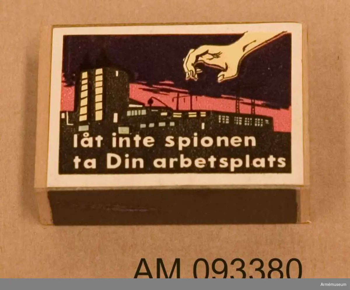 Tändsticksask med tändstickor. På ena sidan motivet Tre stjärnor och på andra sidan ett motiv som illustrerar hur en spionhand sträcker sig över en industribyggnad nattetid. 