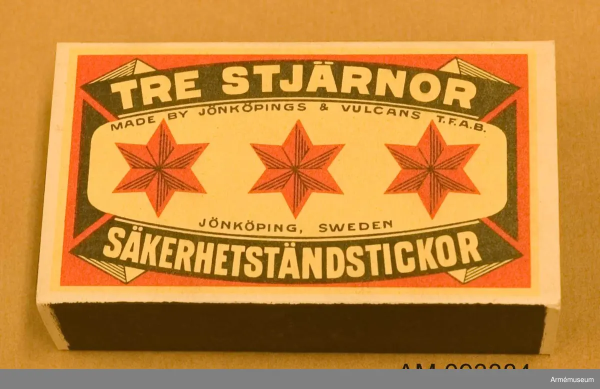 Tändsticksask med tändstickor. På ena sidan motivet Tre stjärnor och på andra sidan ett motiv som illustrerar hur en spionhand sträcker sig över en industribyggnad nattetid.