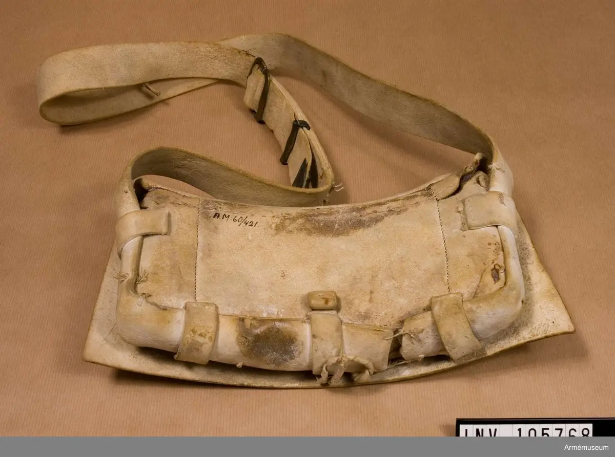 Patronväska av vitt sämskskinn med rem.
Inuti kartuchen finns en bleckplåtsinsats med rum för 12 patroner av ca 18 mm kaliber och plats för en liten oljeflaska. Under locket sitter även en extra ficka,