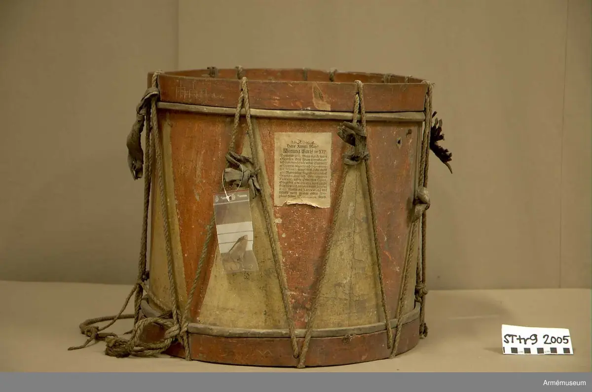 Cylindertrumma av trä med bemålad lim och krita grunderad duk.
Ytan är krackelerad med ett visst bortfall, dock relativt stabil. Tre hål i varierande storlek.

Membran: övre skinnet är smutsigt och repigt med tendenser till sprickbildning. Det finns en centimeterstor lagning. Det undre, har en tvärsgående spricka som tudelar skinnet. Den större delen rullar in mot mitten. Det finns fläckar av ngn. lättlöslig, gråaktig lösning. 

Veckelgjordar: har lindade fogar, den övre med tunt rep/lina, 
Den undre med rep och smalt läderband. Tre tecken inskurna i övre veckelgjordens utsida, som liknar: 1:a,  gaffel med tre klor, 2:a, endast de två yttre klorna och skaft, 3:e tecknet: endast de två yttre klorna, skaftet saknas. 

Spännlina: är fin med tvinnat, avsmalnande repslut. Det finns åtta spännstroppar varav två stycken är fransade.

Spännringar : Utsmyckning : röda kilar målade på gul botten. Mönstret ristat i grunden.

Märkning: fastklistrad Konung Karl den XII med ifyllnad: Litt B  No.4 
Hängandes pappers etikett  i påse No 1211,
Gustavianska koret No 9.