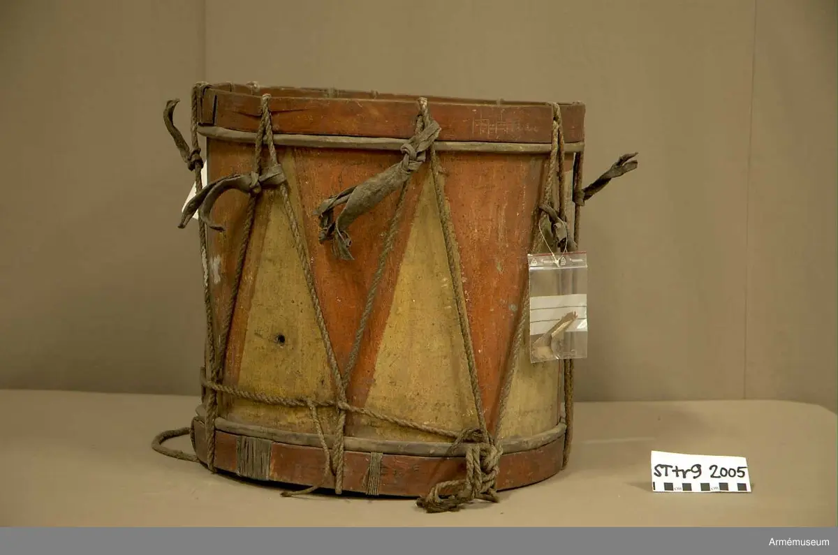 Cylindertrumma av trä med bemålad lim och krita grunderad duk.
Ytan är krackelerad med ett visst bortfall, dock relativt stabil. Tre hål i varierande storlek.

Membran: övre skinnet är smutsigt och repigt med tendenser till sprickbildning. Det finns en centimeterstor lagning. Det undre, har en tvärsgående spricka som tudelar skinnet. Den större delen rullar in mot mitten. Det finns fläckar av ngn. lättlöslig, gråaktig lösning. 

Veckelgjordar: har lindade fogar, den övre med tunt rep/lina, 
Den undre med rep och smalt läderband. Tre tecken inskurna i övre veckelgjordens utsida, som liknar: 1:a,  gaffel med tre klor, 2:a, endast de två yttre klorna och skaft, 3:e tecknet: endast de två yttre klorna, skaftet saknas. 

Spännlina: är fin med tvinnat, avsmalnande repslut. Det finns åtta spännstroppar varav två stycken är fransade.

Spännringar : Utsmyckning : röda kilar målade på gul botten. Mönstret ristat i grunden.

Märkning: fastklistrad Konung Karl den XII med ifyllnad: Litt B  No.4 
Hängandes pappers etikett  i påse No 1211,
Gustavianska koret No 9.