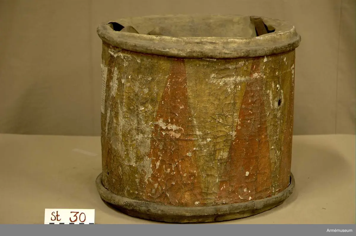 Cylindertrumma av: trä därpå spänd lim o krita grunderad duk som är bemålad. Hål i olika storlek, i jämn rad efter hela sidan. Svårt att säga stick eller insekts hål. Dessutom 7 st. hål i olika storlek, placerade runt om på cylindern. Till höger om den klistrade etiketten finns en stor ruta ca 8x7 cm. med ett totalt bortfall av kredering. För övrigt är ytan mkt. krackelerad och ostabil. 

Membran : övre skinnet är trasigt, undre skinnet är fogat efter spännringen och vågigt.

Veckelgjordar: saknas.

Spännlina: saknas. 

Spännringar : två stycken.

Utsmyckning : bemålad med röda kilar på gul botten. Ristat mönster. 

Märkning : klistrad färdigtryckt Konung Carl den XII etikett med ifyllnad
Litt. B. -No 25.