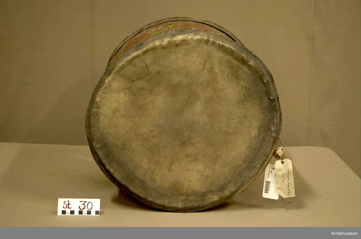 Cylindertrumma av: trä därpå spänd lim o krita grunderad duk som är bemålad. Hål i olika storlek, i jämn rad efter hela sidan. Svårt att säga stick eller insekts hål. Dessutom 7 st. hål i olika storlek, placerade runt om på cylindern. Till höger om den klistrade etiketten finns en stor ruta ca 8x7 cm. med ett totalt bortfall av kredering. För övrigt är ytan mkt. krackelerad och ostabil. 

Membran : övre skinnet är trasigt, undre skinnet är fogat efter spännringen och vågigt.

Veckelgjordar: saknas.

Spännlina: saknas. 

Spännringar : två stycken.

Utsmyckning : bemålad med röda kilar på gul botten. Ristat mönster. 

Märkning : klistrad färdigtryckt Konung Carl den XII etikett med ifyllnad
Litt. B. -No 25.