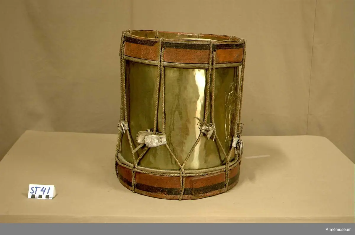 Cylindertrumma av : ljus metall, dekorerad. Metallen är bucklig och har ett mindre hål.

Membran : båda skinnen är trasiga. Ovanpå det övre skinnet ligger en färgad pappskiva. Det undre skinnet har en skada, ca 4 cm. med en flik som går inåt.

Veckelgjordar : träet skiktar sig i den övre veckelgjorden.
 
Spännlina : ser hel ut med 8 st. spännstroppar av vitfärgade tjocka  läderband ca 3 cm. breda. Tre av dessa har tandklippta avslut. 

Spännringar : finns.

Bärring : 

Utsmyckning : vapensköld med mansfigurer på vardera sidan.

Märkning : finns.