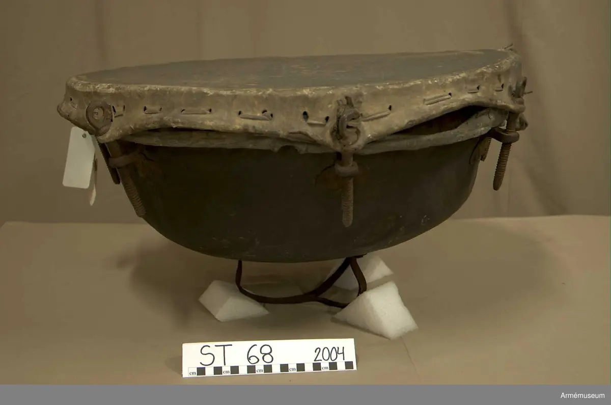 Kittel av koppar och skinn av kalv med fast spännring. Tredelad fot av järn. 7 skruvar och skruvfästen av järn. Bärrem av kalvskinn i två lager med spänne av järn.