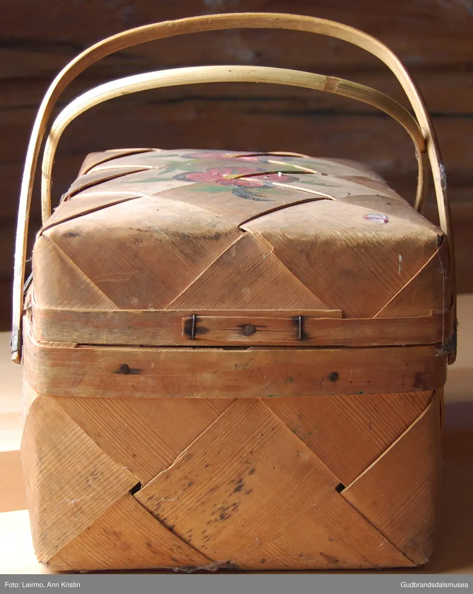 Rektangulær, flettet kurv av furu, håndtak av bambus, to håndtak samles til ett. Med låsemekanisme. Lokk festet til kurven med innslåtte metallkroker.