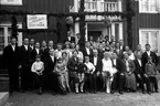 Ett brudpar med släkt och vänner samlade för fotografering av Gustav Andersson, Jönköping, framför en granrisklädd veranda. Där hänger också papperslyktor och en skylt med texten: Lycka och välgång för Olle + Ebba.