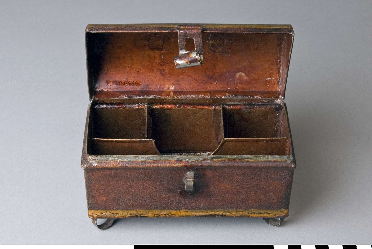 Reseschatull avsett för skrivdon, av bleckplåt. Modell som en liten kista, med kupigt gångjärnsförsett lock och fötter. Inredd med tre fyrkantiga fack och ett smalt som följer framsidan. Endast ett skrivdon finns kvar, en sandströare. På locket ett bygelformat handtag av ståltråd, med två fästen. Fällbar låstunga som passar till en bygel på lådans framsida. Inget lås finns kvar. Fötterna, en i varje hörn, är tillverkade av plåtringar. Schatullet är målat, troligen i rött eller brunt, med gula markeringar runt kanter och handtagsfästen.