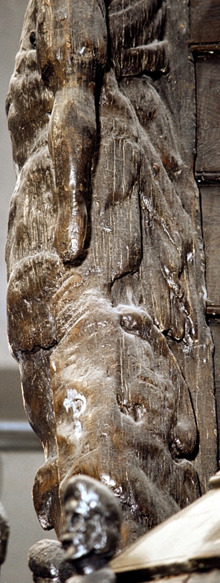 Skulptur i form av en hög, rikt snidad pelare eller stolpe, med kvadratisk genomskärning.

Tre av pelarens sidor är skulpterade medan den fjärde utgör anliggningsyta, försedd med ett antal olikformade urtag. De skulpterade sidorna är horisontellt indelade i tre eller fyra sektioner eller nivåer, avgränsade från varandra av profilerade och snedställda vulster. Nedtill avslutas skulpturen av en ornerad och på mitten insvängd formation.



Ovanför denna nedre avslutning är pelaren utformad som en reslig mansfigur, vänd mot vänster, sedd från betraktaren. Mansfiguren, som täcker tre av skulpturens fyra sidor, är iförd en romersk rustning med en bred fältbindel draperad från höger axel snett över bröstet. På huvudet med det lockiga och utslagna håret sitter en krona av öppen typ, med liljeliknande ornament. Figurens vänstra arm är böjd, medan den högra armen är sträckt utmed sidan. Handen till den högra armen saknas, men har hållit en rikt profilerad sköld, med mycket slitna och vaga konturer av en på bakbenen mot vänster gående grip med stora vingar. Skölden är snidad i ett med figuren, men med kraftiga underskärningar. Vid figurens fötter skymtar ett djur av något slag. Bakom mannens huvud tycks ett snidat textilstycke, kanske del till ett draperi, vara anbragt.



Ovanför den romerska krigaren eller riddaren har pelaren formen av en herm utan armar med långt, krusigt hår och mustaschprytt ansikte. Liksom mansfiguren täcker även hermen tre sidor av pelaren. Huvudet är vridet mot vänster. Överkroppen är bar så när som på ett flikigt plagg som täcker axlar och övre delen av bröstet. Från huvudet och runt midjan är ett tygstycke draperat. Hermens nederdel är formad till en stor maskaron med uppspärrad mun och utsträckt tunga. Maskaronen övergår upptill och utmed sidorna i yppiga bladformationer.



Pelaren avslutas upptill av ett knoppliknande ornament. Ornamentet är ornerat med akantusbladliknande bildningar samt fyra byglar som möts på toppen, där vissa spår antyder att det ursprungligen suttit ett klot eller dylikt, men som fallit bort. Eventuellt kan ornamentet utgöra en huvudbonad till hermen.

Skulpturen är kraftigt sliten.



Text in English: Sculpture in the shape of a richly carved column or pillar.

Three sides of the pillar are carved, while the fourth side comprises the attachment surface and has a number of variously-shaped indentations. The sculptured sides are divided horizontally into three or four sections separated by angled tori.



The lower section of the pillar is carved in the figure of a tall man. The figure is clad in Roman armour and has a field badge (cloak/plaid) draped across his chest at an angle from his right shoulder. On his head he wears an open crown. He has long, curly hair. The right arm is broken off, the left is at his side. The left hand is missing but once held a richly carved shield.

The shield is carved out of the same piece of wood as the figure, but deeply undercut. A carved figure is apparent on the shield. Although the shield is badly worn, it is possible to recognise one of the hind legs of a griffin with large wings. At the man's feet lies an animal of some kind.



Above the Roman warrior or knight, the pillar is carved in the shape of a herm, without arms and with long, curly hair and a moustache. Like the male figure, the herm also covers three sides of the pillar. The head is twisted to the left. The upper part of the body is bare except for a piece of cloth that covers the shoulders and upper chest. The lower part of the herm is carved as a large mask, its mouth open and tongue protruding. The sides and upper parts of the mask are carved as luxuriant leaves.



The upper part of the pillar has a ornament in shape of a knob. The ornament has a ring of acanthus leaves and four spherical loops run vertically from the acanthus leaves and meet at a point at the top. An object, for example a globe appears to have adorned the top of the ornament. This object is missing. The ornament may also be a part of the headgear of the herm.

The sculpture is severely worn.