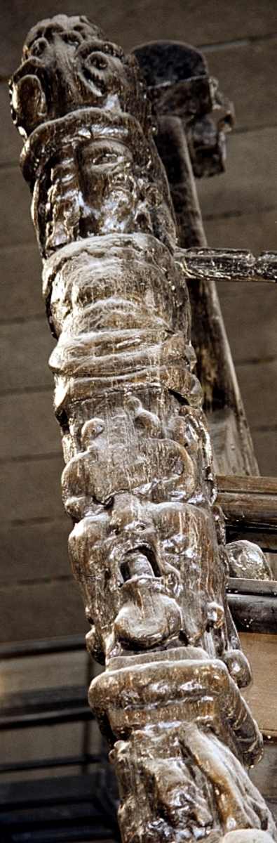Skulptur i form av en hög, rikt snidad pelare eller stolpe, med kvadratisk genomskärning.

Tre av pelarens sidor är skulpterade medan den fjärde utgör anliggningsyta, försedd med ett antal olikformade urtag. De skulpterade sidorna är horisontellt indelade i tre eller fyra sektioner eller nivåer, avgränsade från varandra av profilerade och snedställda vulster. Nedtill avslutas skulpturen av en ornerad och på mitten insvängd formation.



Ovanför denna nedre avslutning är pelaren utformad som en reslig mansfigur, vänd mot vänster, sedd från betraktaren. Mansfiguren, som täcker tre av skulpturens fyra sidor, är iförd en romersk rustning med en bred fältbindel draperad från höger axel snett över bröstet. På huvudet med det lockiga och utslagna håret sitter en krona av öppen typ, med liljeliknande ornament. Figurens vänstra arm är böjd, medan den högra armen är sträckt utmed sidan. Handen till den högra armen saknas, men har hållit en rikt profilerad sköld, med mycket slitna och vaga konturer av en på bakbenen mot vänster gående grip med stora vingar. Skölden är snidad i ett med figuren, men med kraftiga underskärningar. Vid figurens fötter skymtar ett djur av något slag. Bakom mannens huvud tycks ett snidat textilstycke, kanske del till ett draperi, vara anbragt.



Ovanför den romerska krigaren eller riddaren har pelaren formen av en herm utan armar med långt, krusigt hår och mustaschprytt ansikte. Liksom mansfiguren täcker även hermen tre sidor av pelaren. Huvudet är vridet mot vänster. Överkroppen är bar så när som på ett flikigt plagg som täcker axlar och övre delen av bröstet. Från huvudet och runt midjan är ett tygstycke draperat. Hermens nederdel är formad till en stor maskaron med uppspärrad mun och utsträckt tunga. Maskaronen övergår upptill och utmed sidorna i yppiga bladformationer.



Pelaren avslutas upptill av ett knoppliknande ornament. Ornamentet är ornerat med akantusbladliknande bildningar samt fyra byglar som möts på toppen, där vissa spår antyder att det ursprungligen suttit ett klot eller dylikt, men som fallit bort. Eventuellt kan ornamentet utgöra en huvudbonad till hermen.

Skulpturen är kraftigt sliten.



Text in English: Sculpture in the shape of a richly carved column or pillar.

Three sides of the pillar are carved, while the fourth side comprises the attachment surface and has a number of variously-shaped indentations. The sculptured sides are divided horizontally into three or four sections separated by angled tori.



The lower section of the pillar is carved in the figure of a tall man. The figure is clad in Roman armour and has a field badge (cloak/plaid) draped across his chest at an angle from his right shoulder. On his head he wears an open crown. He has long, curly hair. The right arm is broken off, the left is at his side. The left hand is missing but once held a richly carved shield.

The shield is carved out of the same piece of wood as the figure, but deeply undercut. A carved figure is apparent on the shield. Although the shield is badly worn, it is possible to recognise one of the hind legs of a griffin with large wings. At the man's feet lies an animal of some kind.



Above the Roman warrior or knight, the pillar is carved in the shape of a herm, without arms and with long, curly hair and a moustache. Like the male figure, the herm also covers three sides of the pillar. The head is twisted to the left. The upper part of the body is bare except for a piece of cloth that covers the shoulders and upper chest. The lower part of the herm is carved as a large mask, its mouth open and tongue protruding. The sides and upper parts of the mask are carved as luxuriant leaves.



The upper part of the pillar has a ornament in shape of a knob. The ornament has a ring of acanthus leaves and four spherical loops run vertically from the acanthus leaves and meet at a point at the top. An object, for example a globe appears to have adorned the top of the ornament. This object is missing. The ornament may also be a part of the headgear of the herm.

The sculpture is severely worn.