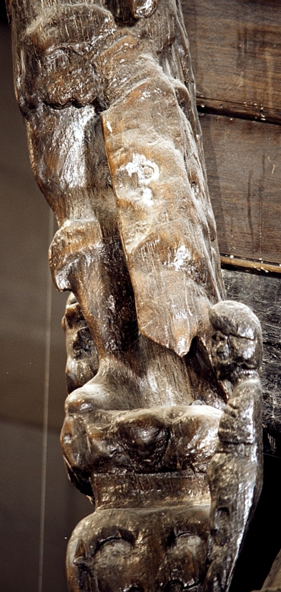 Skulptur i form av en hög, rikt snidad pelare eller stolpe, med kvadratisk genomskärning.

Tre av pelarens sidor är skulpterade medan den fjärde utgör anliggningsyta, försedd med ett antal olikformade urtag. De skulpterade sidorna är horisontellt indelade i tre eller fyra sektioner eller nivåer, avgränsade från varandra av profilerade och snedställda vulster. Nedtill avslutas skulpturen av en ornerad och på mitten insvängd formation.



Ovanför denna nedre avslutning är pelaren utformad som en reslig mansfigur, vänd mot vänster, sedd från betraktaren. Mansfiguren, som täcker tre av skulpturens fyra sidor, är iförd en romersk rustning med en bred fältbindel draperad från höger axel snett över bröstet. På huvudet med det lockiga och utslagna håret sitter en krona av öppen typ, med liljeliknande ornament. Figurens vänstra arm är böjd, medan den högra armen är sträckt utmed sidan. Handen till den högra armen saknas, men har hållit en rikt profilerad sköld, med mycket slitna och vaga konturer av en på bakbenen mot vänster gående grip med stora vingar. Skölden är snidad i ett med figuren, men med kraftiga underskärningar. Vid figurens fötter skymtar ett djur av något slag. Bakom mannens huvud tycks ett snidat textilstycke, kanske del till ett draperi, vara anbragt.



Ovanför den romerska krigaren eller riddaren har pelaren formen av en herm utan armar med långt, krusigt hår och mustaschprytt ansikte. Liksom mansfiguren täcker även hermen tre sidor av pelaren. Huvudet är vridet mot vänster. Överkroppen är bar så när som på ett flikigt plagg som täcker axlar och övre delen av bröstet. Från huvudet och runt midjan är ett tygstycke draperat. Hermens nederdel är formad till en stor maskaron med uppspärrad mun och utsträckt tunga. Maskaronen övergår upptill och utmed sidorna i yppiga bladformationer.



Pelaren avslutas upptill av ett knoppliknande ornament. Ornamentet är ornerat med akantusbladliknande bildningar samt fyra byglar som möts på toppen, där vissa spår antyder att det ursprungligen suttit ett klot eller dylikt, men som fallit bort. Eventuellt kan ornamentet utgöra en huvudbonad till hermen.

Skulpturen är kraftigt sliten.



Text in English: Sculpture in the shape of a richly carved column or pillar.

Three sides of the pillar are carved, while the fourth side comprises the attachment surface and has a number of variously-shaped indentations. The sculptured sides are divided horizontally into three or four sections separated by angled tori.



The lower section of the pillar is carved in the figure of a tall man. The figure is clad in Roman armour and has a field badge (cloak/plaid) draped across his chest at an angle from his right shoulder. On his head he wears an open crown. He has long, curly hair. The right arm is broken off, the left is at his side. The left hand is missing but once held a richly carved shield.

The shield is carved out of the same piece of wood as the figure, but deeply undercut. A carved figure is apparent on the shield. Although the shield is badly worn, it is possible to recognise one of the hind legs of a griffin with large wings. At the man's feet lies an animal of some kind.



Above the Roman warrior or knight, the pillar is carved in the shape of a herm, without arms and with long, curly hair and a moustache. Like the male figure, the herm also covers three sides of the pillar. The head is twisted to the left. The upper part of the body is bare except for a piece of cloth that covers the shoulders and upper chest. The lower part of the herm is carved as a large mask, its mouth open and tongue protruding. The sides and upper parts of the mask are carved as luxuriant leaves.



The upper part of the pillar has a ornament in shape of a knob. The ornament has a ring of acanthus leaves and four spherical loops run vertically from the acanthus leaves and meet at a point at the top. An object, for example a globe appears to have adorned the top of the ornament. This object is missing. The ornament may also be a part of the headgear of the herm.

The sculpture is severely worn.