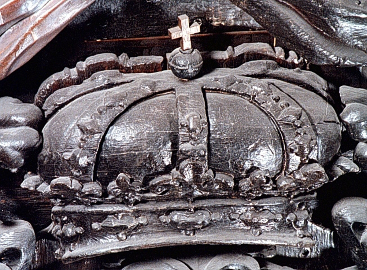 Skulpterad, kunglig krona. Undersidan är perspektiviskt framställd genom att kronans baksida gjorts högre än framsidan. Kronringen är upptill prydd med omväxlande större och mindre liljeliknande ornament. På själva ringen och på byglarna sitter fasettslipade "ädelstenar". Kronans baksida är slät.
Kronan är välbevarad.

Text in English: Royal crown. The underside is carved in perspective, achieved by carving the back of the crown higher than the front. The circlet is decorated at the top with lily-like ornaments of varying size. On the circlet itself and on the verticals are faceted "precious stones". The reverse side of the crown is smooth.
The sculpture is well preserved.