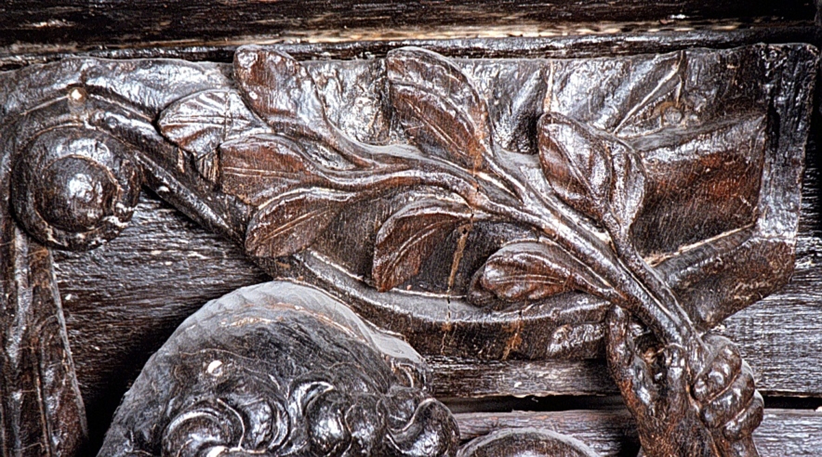 Skulpterad putto figur stående på postament med omgivande ornamentik.
Putton har ett frodigt och undersätsigt utseende, med avrundade former. Puttons vänstra hand vilar på en sköld, medan höger hand är uppåtsträckt, hållande en olivkvist.
Ovanför putton sitter ett veckrikt draperi, "upphängt" på en stång med profilerad ände. Där det nedhängande draperiet slutar vidtar tunga fruktfestoner. Festonerna når ned till postamentet, som är dekorerat med symmetriskt ställda rullverksspiraler. Rullverksspiralerna är utformade så att de bildar krumma vädurs- eller gumsehorn till ett i postamentet utsnidat djurhuvud. Skulpturens baksida är slät.
Puttons ansikte, vänster arm samt mage är snidade separat.

Text in English: Sculpture of a putto, standing on a base surrounded by ornamentation.
The exuberant, plump putto has a rounded body. The putto''s left hand rests on a shield. The right hand is holding an olive branch aloft. Above the putto''s head hangs a heavily-folded drapery.
The drapery forms a swag as it hangs from a curtain rod. The prominent ends of the curtain rod are carved in relief. Heavy festoons of fruit flow from the drapery ends to the feet of the putto. The symmetrical scrollwork on the base takes on the appearance of ram''s horns when combined with the imprecise representations of animal heads. The sculpture''s reverse side is smooth.
Face, right arm and belly are carved separately.