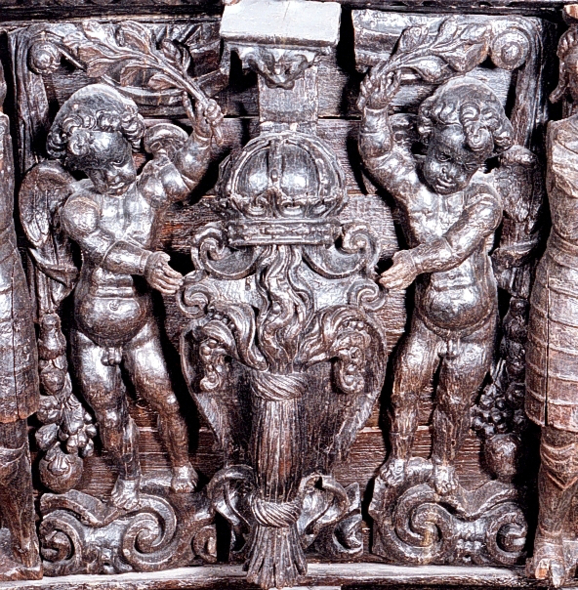 Skulpterad putto figur stående på postament med omgivande ornamentik.
Putton har ett frodigt och undersätsigt utseende, med avrundade former. Puttons vänstra hand vilar på en sköld, medan höger hand är uppåtsträckt, hållande en olivkvist.
Ovanför putton sitter ett veckrikt draperi, "upphängt" på en stång med profilerad ände. Där det nedhängande draperiet slutar vidtar tunga fruktfestoner. Festonerna når ned till postamentet, som är dekorerat med symmetriskt ställda rullverksspiraler. Rullverksspiralerna är utformade så att de bildar krumma vädurs- eller gumsehorn till ett i postamentet utsnidat djurhuvud. Skulpturens baksida är slät.
Puttons ansikte, vänster arm samt mage är snidade separat.

Text in English: Sculpture of a putto, standing on a base surrounded by ornamentation.
The exuberant, plump putto has a rounded body. The putto''s left hand rests on a shield. The right hand is holding an olive branch aloft. Above the putto''s head hangs a heavily-folded drapery.
The drapery forms a swag as it hangs from a curtain rod. The prominent ends of the curtain rod are carved in relief. Heavy festoons of fruit flow from the drapery ends to the feet of the putto. The symmetrical scrollwork on the base takes on the appearance of ram''s horns when combined with the imprecise representations of animal heads. The sculpture''s reverse side is smooth.
Face, right arm and belly are carved separately.