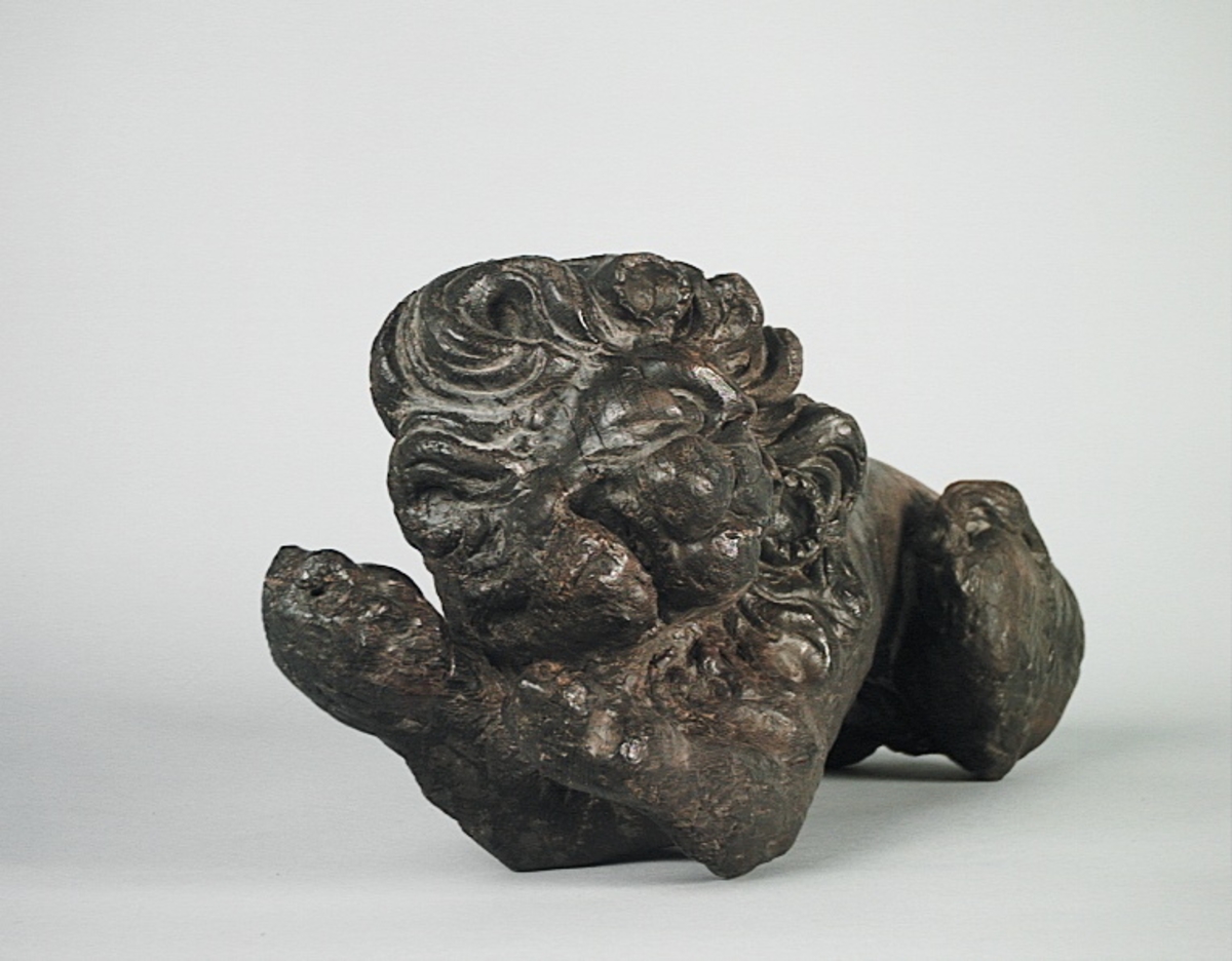 Skulptur av ett litet, liggande lejon med utsträckta framben och hopdragna bakben.
Lejonet är riktat mot vänster, sett från betraktaren. Det snett hållna huvudet återges i det närmaste i en face. Större delen av svansen saknas. Den befintliga delen av svansen avtecknar sig mot lejonets vänstra lår.

Lejonet är endast delvist rundskulpterat. På baksidan, i höjd med huvudet och höger framben, återfinns ett stort, snett urtag som bildar en slät anliggningsyta. Skulpturens framparti är således endast till hälften så tjockt som dess bakparti.
Bakkroppen är kraftigt sliten och skadad. Vänster bakben saknas, liksom större delen av svansen.

Text in English: Sculpture of a small reclining lion with its forelegs extended and its  hind legs tucked beneath its body.
Facing to the observer''s left, the head, held crookedly, is nearly en face. A large part of the tail is missing, and the remainder curves towards the left hind leg.

The lion is only partly three-dimensional. On the rear at the head height and at the right foreleg a large oblique hole forms a flat area for attachement, thus the forepart of the sculpture is thus only half as thick as its rear.
The rear of the body is badly worn and damaged. The left hind leg has been broken off.