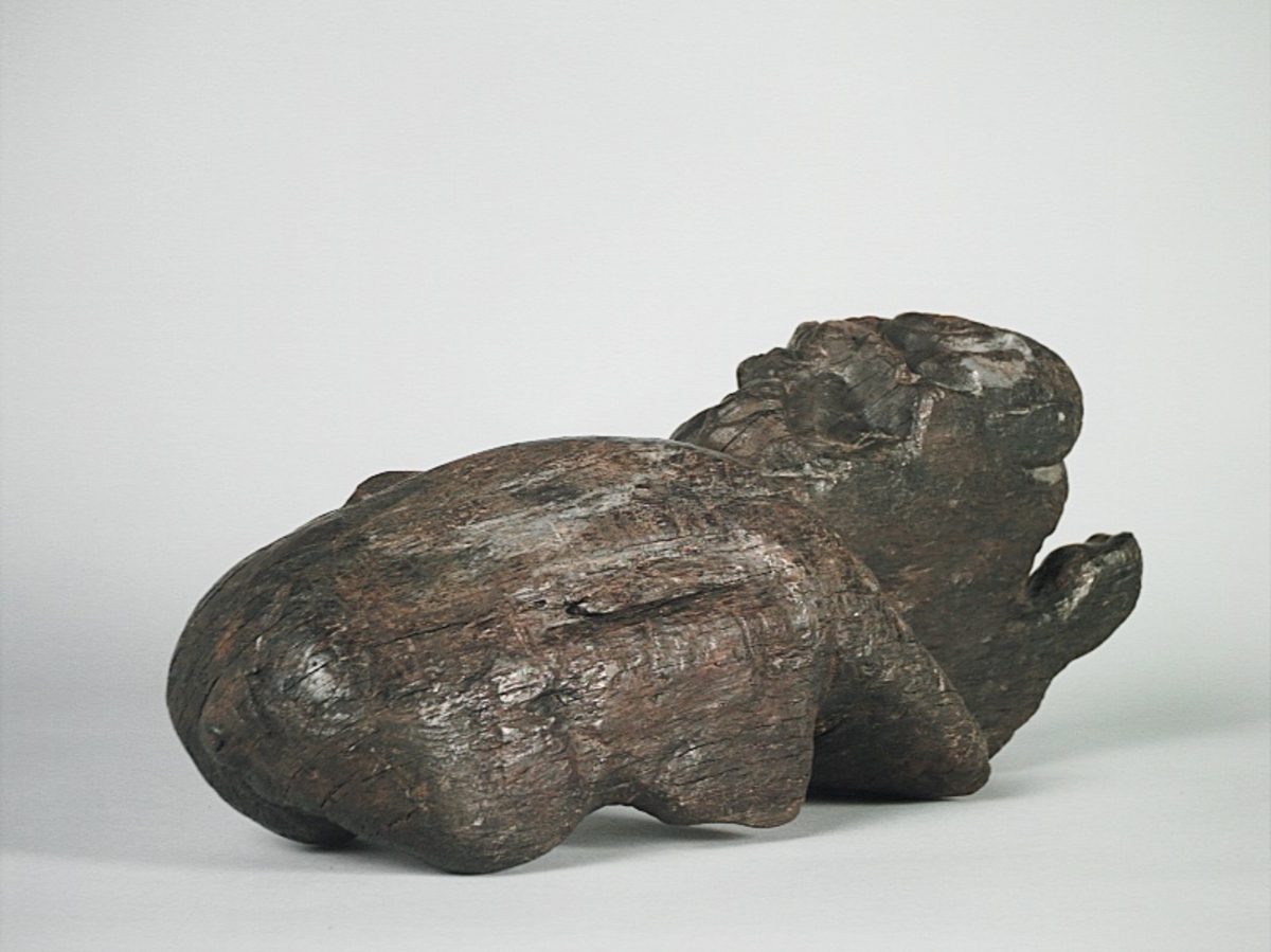 Skulptur av ett litet, liggande lejon med utsträckta framben och hopdragna bakben.
Lejonet är riktat mot vänster, sett från betraktaren. Det snett hållna huvudet återges i det närmaste i en face. Större delen av svansen saknas. Den befintliga delen av svansen avtecknar sig mot lejonets vänstra lår.

Lejonet är endast delvist rundskulpterat. På baksidan, i höjd med huvudet och höger framben, återfinns ett stort, snett urtag som bildar en slät anliggningsyta. Skulpturens framparti är således endast till hälften så tjockt som dess bakparti.
Bakkroppen är kraftigt sliten och skadad. Vänster bakben saknas, liksom större delen av svansen.

Text in English: Sculpture of a small reclining lion with its forelegs extended and its  hind legs tucked beneath its body.
Facing to the observer''s left, the head, held crookedly, is nearly en face. A large part of the tail is missing, and the remainder curves towards the left hind leg.

The lion is only partly three-dimensional. On the rear at the head height and at the right foreleg a large oblique hole forms a flat area for attachement, thus the forepart of the sculpture is thus only half as thick as its rear.
The rear of the body is badly worn and damaged. The left hind leg has been broken off.